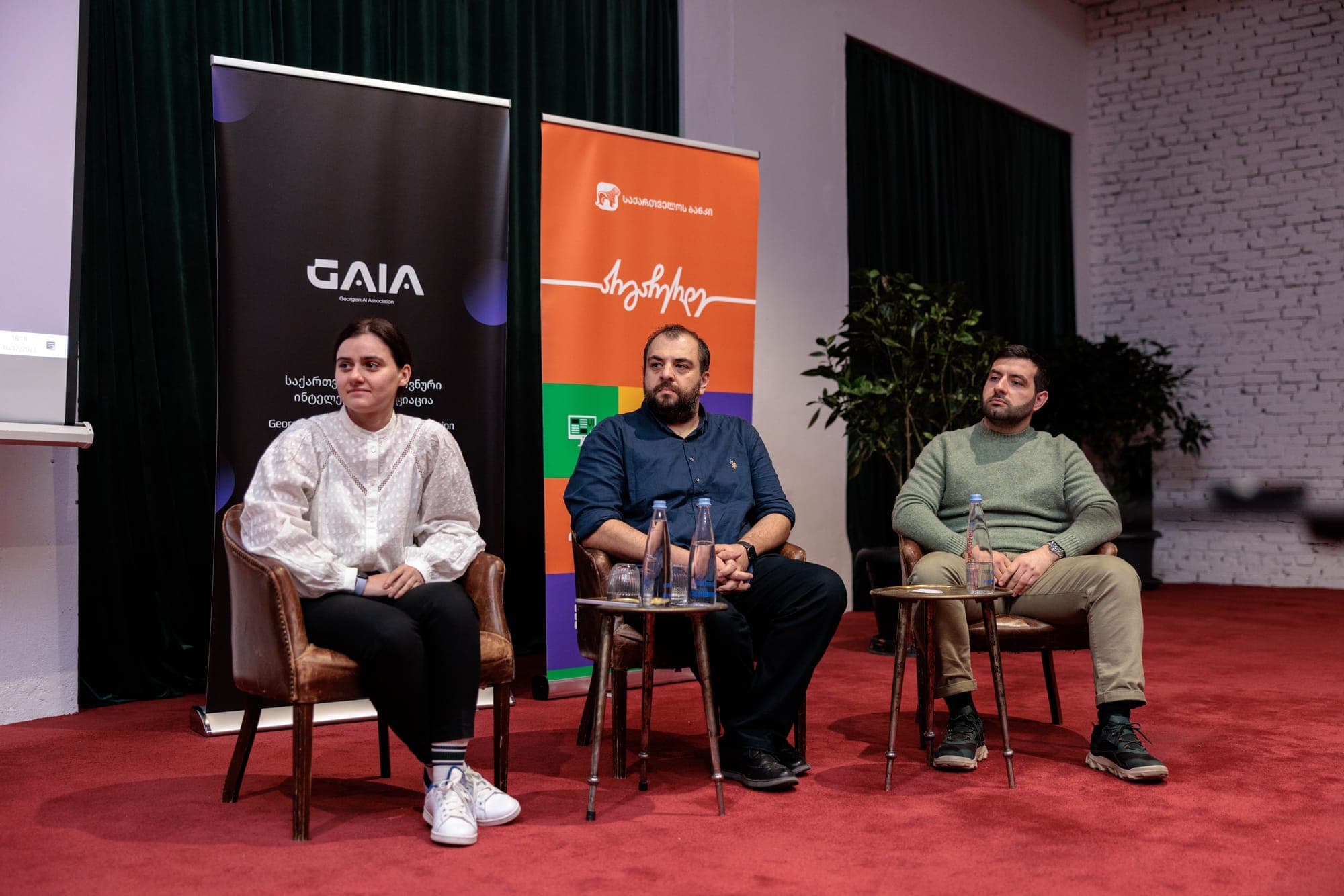 პანელური დისკუსია GAIA-ის ბანერის წინ: სამი მონაწილე სავარძლებში